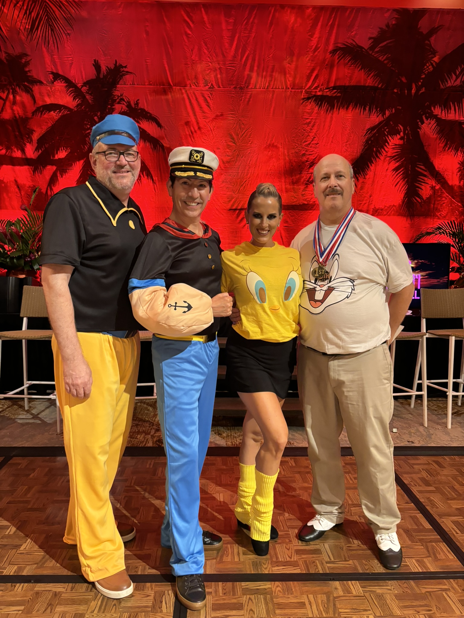 Amy Hanson in a yellow cartoon-bird costume with three fellow dancers in cartoon-themed costumes in front of a red palm-tree backdrop, including her student Jerry wearing a competition medal