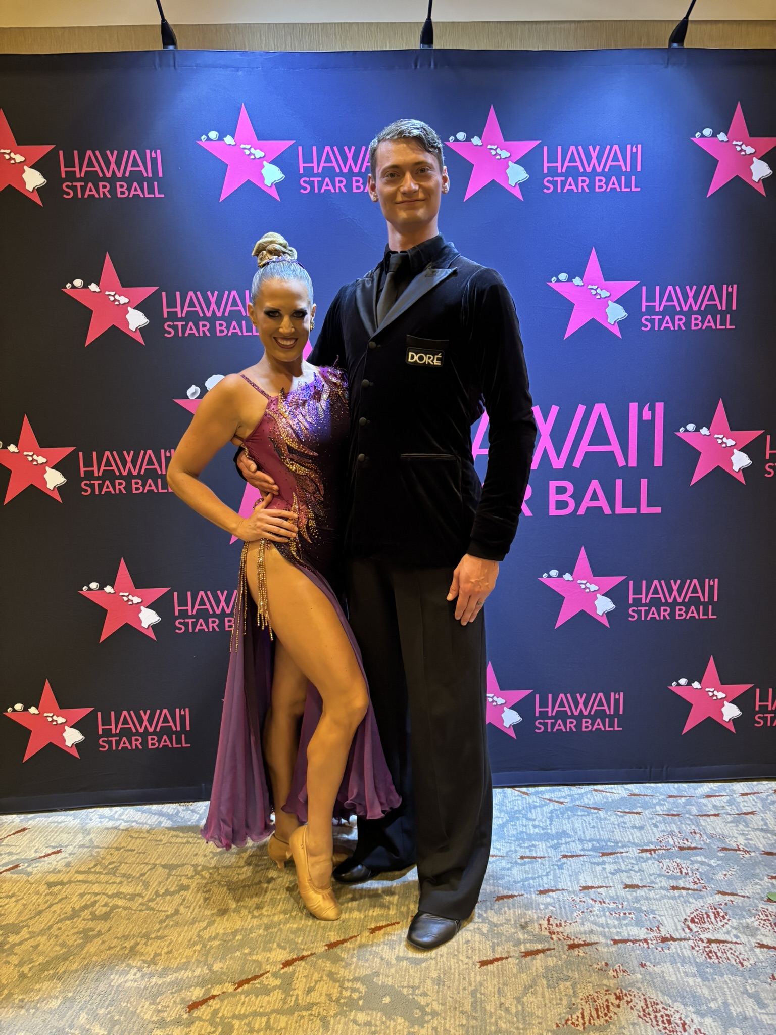 Amy Hanson in a purple competition gown standing with her professional partner in a black velvet tailsuit in front of the pink-starred Hawaii Star Ball backdrop