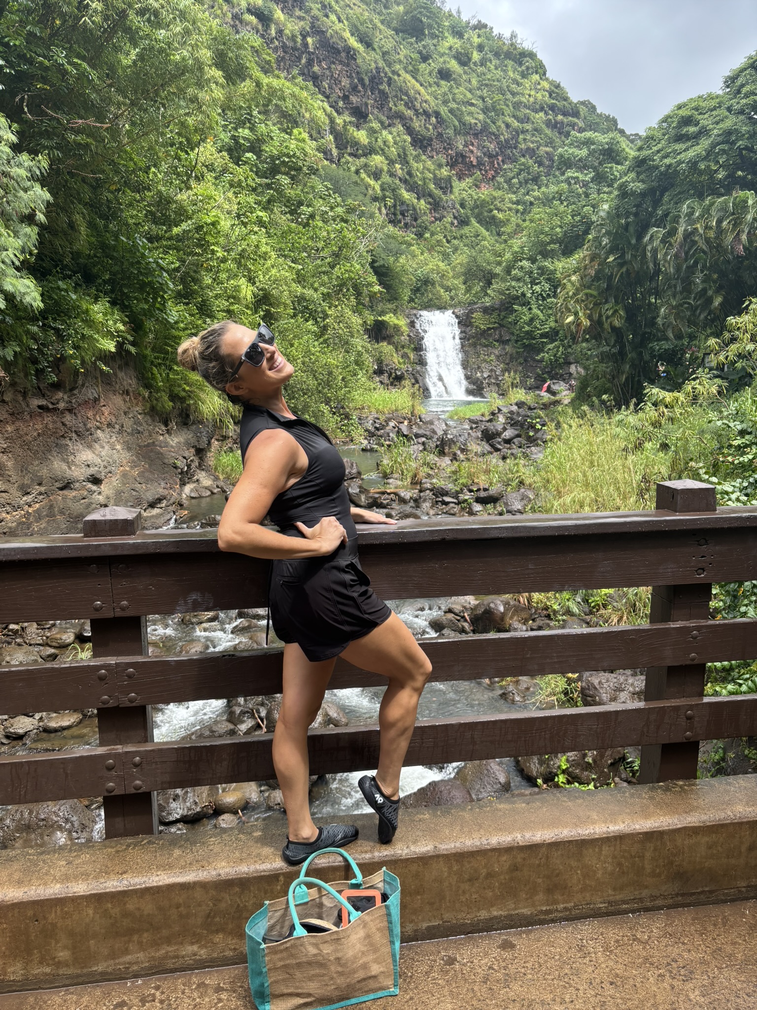 Amy Hanson smiling and leaning on a wooden bridge railing with Waimea Falls and lush green cliffs behind her on Oahu's North Shore