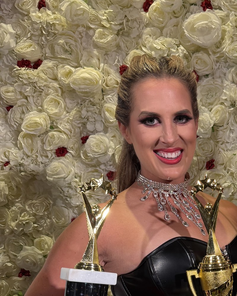 Amy Hanson of Ballroom by Amy smiling with two trophies in front of a white flower wall
