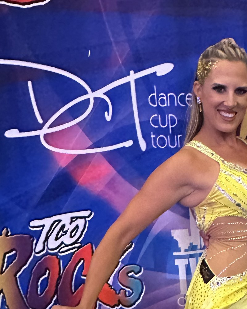 Amy Hanson of Ballroom by Amy in a yellow ballroom gown in front of a Dancer’s Cup Tour backdrop