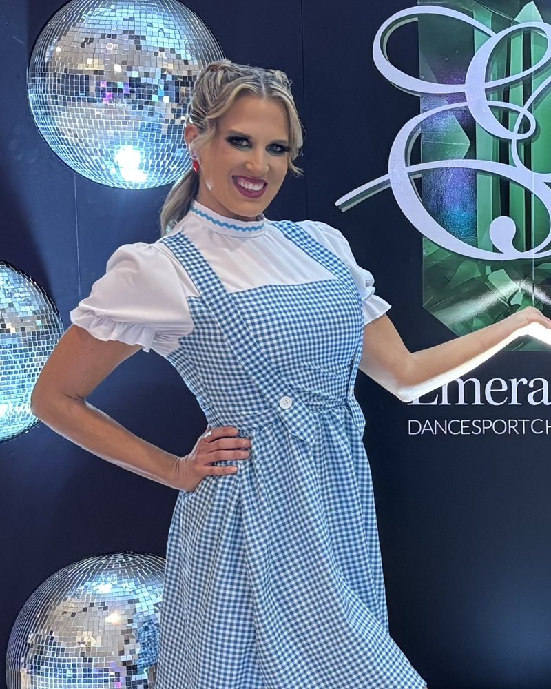 Amy Hanson of Ballroom by Amy smiling in a blue gingham theme costume in front of mirror balls at a ballroom competition
