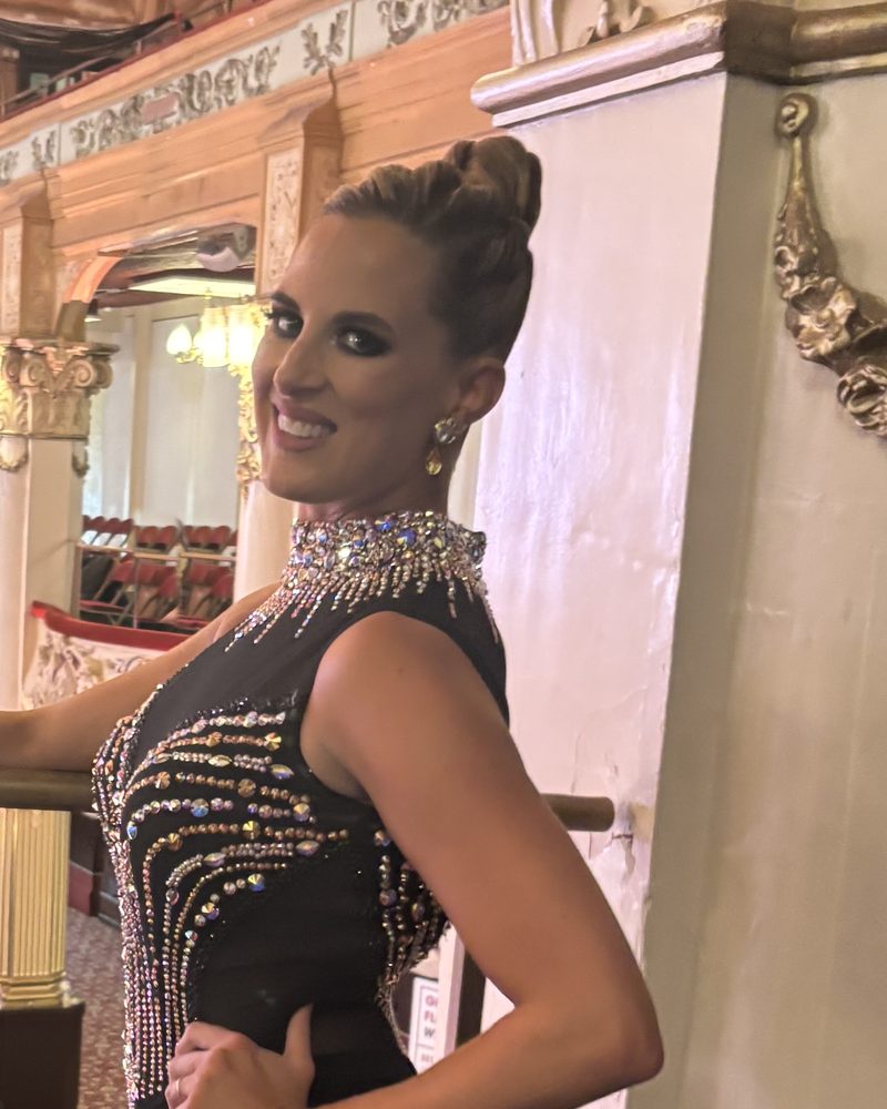 Amy Hanson of Ballroom by Amy smiling in a black rhinestone ballroom gown on a theater balcony