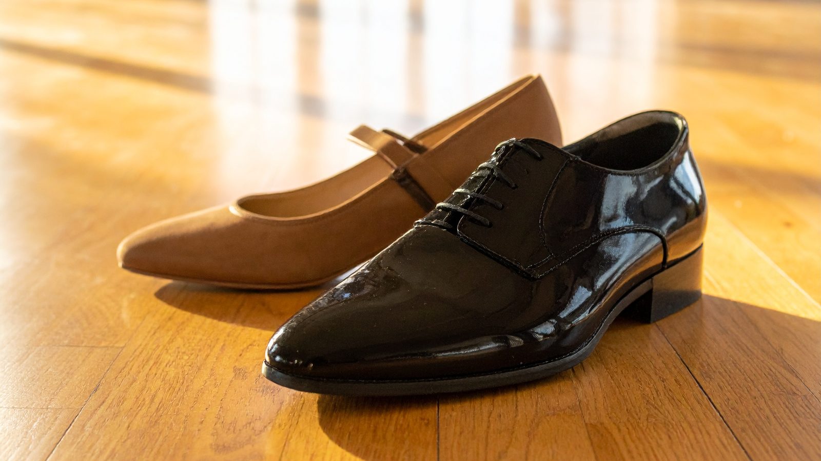Illustration: a man's black patent ballroom shoe beside a woman's tan strapped ballroom shoe on a sunlit wooden ballroom floor