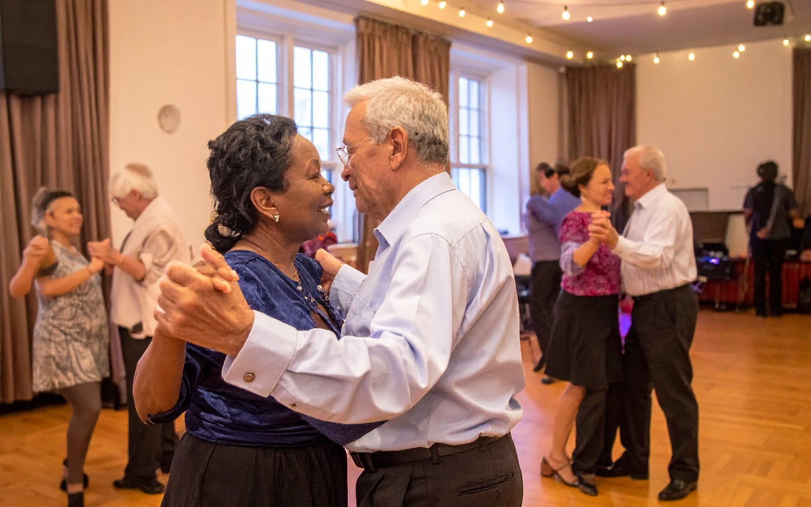 Illustration of couples of different ages dancing a foxtrot at a social dance night in a sunlit hall with string lights